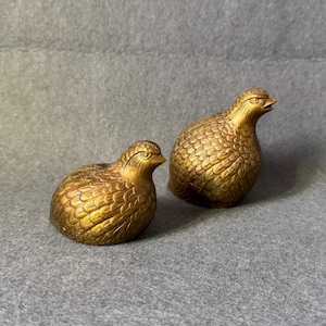 May include: Two golden-colored bird figurines. The birds have detailed feather textures and are posed in a resting position. The figurines are on a gray surface.