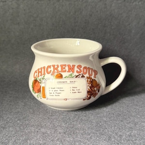 Op de afbeelding: Crèmekleurige keramische soepmok met een handvat, met de woorden "CHICKEN SOUP" in oranje. De mok bevat een recept met ingrediënten zoals kip, water en kruiden. Er zijn ook decoratieve illustraties van groenten en kruiden.
