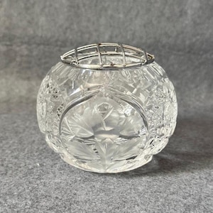 Könnte beinhalten: Eine klare, runde Kristall-Blumenvase mit detailliertem Schliffmuster. Die Vase hat ein silbernes Metallgitter oben zur Anordnung von Blumen. Das Design beinhaltet ein Blumenmotiv, das dem Stück einen Hauch von Eleganz verleiht.