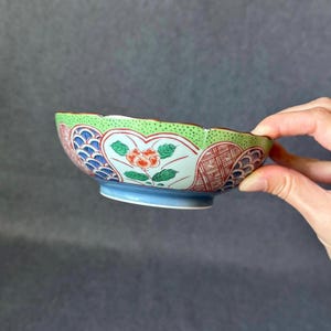 May include: A small, decorative ceramic bowl with a scalloped edge. The bowl features a floral design with red, green, and blue accents. The base is light blue, and the rim is green. The bowl is held by a hand.