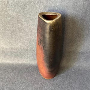 Könnte beinhalten: Eine hohe Keramikvase mit dreieckiger Öffnung. Die Vase hat einen Farbverlauf, der von einem rötlich-braunen Farbton am Boden zu einem dunkelbraunen und goldenen Farbton an der Oberseite übergeht. Die Vase hat eine glatte, polierte Oberfläche.