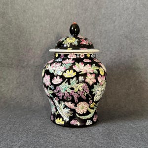 May include: A black ceramic ginger jar with a matching lid featuring a small black finial. The surface is decorated with a colorful floral pattern including pink, yellow, and purple flowers with green leaves covering the entire exterior.