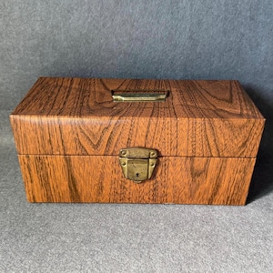 May include: A rectangular wooden box with a faux wood grain finish. The box has a brass-colored latch and handle. The box is closed and sits on a gray surface.