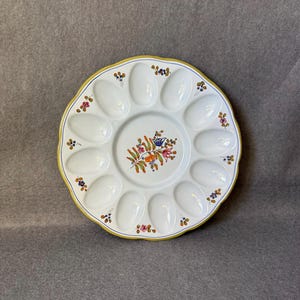 以下が含まれることがあります： 12個の卵型のくぼみがある白いセラミック製の卵用サービングプラッター。プラッターは、黄色の縁と青い内側の境界線があり、中央と縁の周りに花の模様で装飾されています。