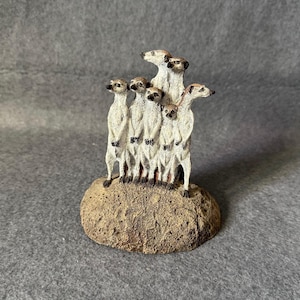 Könnte beinhalten: Eine Figur einer Gruppe von Erdmännchen, die zusammen auf einem kleinen, strukturierten, braunen Felsen stehen. Die Erdmännchen haben hellbraunes und braunes Fell, dunkle Augen und eine aufrechte Haltung. Der Hintergrund ist einfarbig grau.