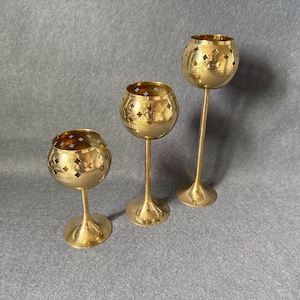 May include: Three gold-colored candle holders with a decorative pierced design. Each holder has a round bowl shape with a long stem and a circular base. The candle holders vary in height, creating a tiered display.