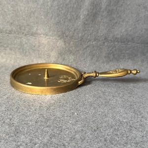 May include: A vintage brass candle holder with a round base and a handle. The candle holder has a central spike for securing a candle and decorative scrollwork. The handle is ornate, adding to the antique aesthetic. The overall design is simple yet elegant.