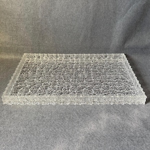 May include: A clear, rectangular decorative tray with a textured surface and a scalloped edge. The tray is made of a transparent material and sits on a grey surface. The tray is a decorative item.