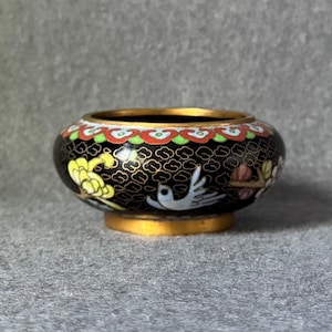 Op de afbeelding: Een kleine, ronde, decoratieve kom met een zwarte achtergrond en gouden rand. De kom heeft kleurrijke bloemen- en vogelontwerpen. De basis is goudkleurig en de rand is afgewerkt met rode, groene en witte accenten.