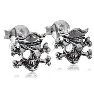 Op de afbeelding: Een paar zilverkleurige oorstekers, elk in de vorm van een piraten schedel met een hoed, ooglapje en gekruiste botten. De oorbellen hebben een gedetailleerde, getextureerde afwerking en een steker sluiting. Het metaal is gemarkeerd met '925'.