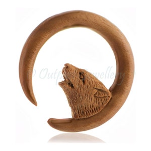 May include: A brown wooden ear stretcher featuring a carved wolf head. The stretcher is a circular shape, with the wolf head carved on the side. The wolf is shown with its mouth open, as if howling.