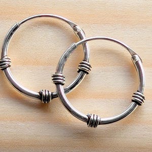 以下が含まれることがあります： 銀色のフープピアスのペア。各イヤリングは、間隔をあけて装飾的なワイヤーラッピングが施された円形デザインです。イヤリングは、明るい茶色の木製の表面に表示されています。