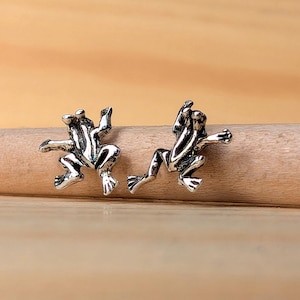 May include: A pair of silver-toned frog stud earrings. Each earring depicts a frog in a leaping pose, with outstretched limbs and detailed features. The earrings are set against a light brown wooden background.