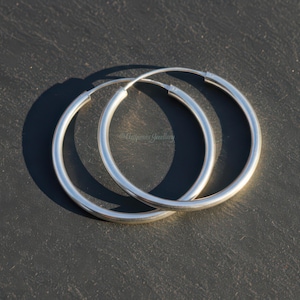 Op de afbeelding: Een paar zilveren oorringen. De oorbellen zijn cirkelvormig en hebben een glad, gepolijst oppervlak. De oorringen worden getoond tegen een donkere, getextureerde achtergrond, met een schaduw.