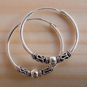 Puede incluir: Un par de pendientes de aro plateados con detalles decorativos. Cada pendiente presenta una serie de adornos oscuros y una cuenta central plateada. Los pendientes se muestran sobre una superficie de madera marrón claro.