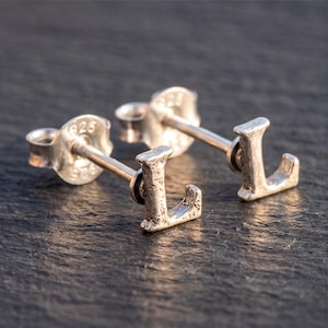 May include: A pair of sterling silver stud earrings shaped like the letter L. Each earring features a textured, rustic finish and includes a standard post back with a 925 hallmark stamped on the circular backing, indicating silver composition.