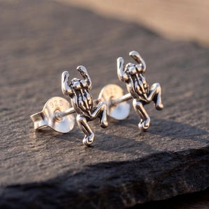 Può includere: Un paio di orecchini a lobo in argento a forma di piccole rane con braccia e gambe sollevate. Il metallo ha una finitura lucida con dettagli ossidati scuri per evidenziare i tratti anatomici. Ogni orecchino presenta il marchio 925 sul retro.