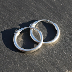 May include: A pair of silver hoop earrings. The earrings are circular, with a smooth, polished surface. They are displayed on a dark, textured surface, with sunlight casting shadows. The earrings are a classic jewelry item.
