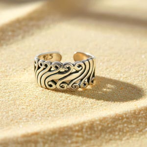Può includere: Un anello regolabile color argento con un design aperto. L'anello presenta un intricato motivo di linee e riccioli a spirale. Il numero "925" è stampato sull'anello. L'anello è esposto su una superficie sabbiosa.