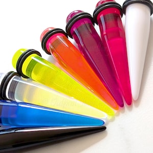 May include: A selection of brightly coloured ear gauges in shades of black, blue, yellow, orange, pink, and white. Each gauge has a tapered design and is made of a translucent material. The gauges are arranged in a fan shape.