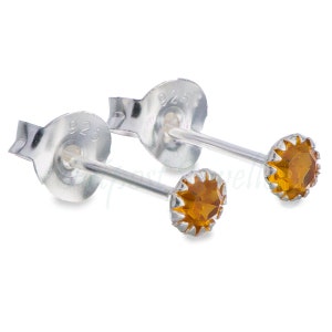 May include: A pair of sterling silver stud earrings with amber-colored gemstones. The earrings have a small, round gemstone set in a scalloped bezel. The posts and backs are also silver, with a 925 stamp.