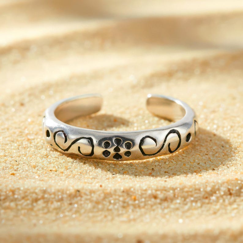 Peut inclure: Bague d'orteil r&eacute;glable argent&eacute;e orn&eacute;e d'un motif de volutes et de points. La bague est ouverte en haut et repose sur un fond de sable.