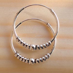 Op de afbeelding: Een paar zilveren oorringen. Elke oorring heeft een reeks kleine, getextureerde kralen en een grotere, gladde kraal. De oorbellen zijn geplaatst op een lichtbruin houten oppervlak.