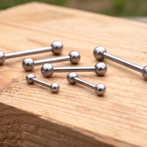 May include: A selection of silver-coloured barbell body jewellery. The barbells feature a straight bar with a ball at each end. They are displayed on a light brown wooden surface.