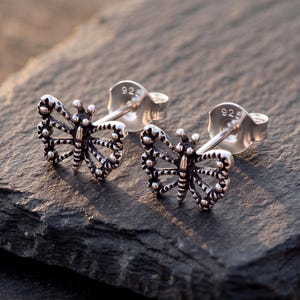 Könnte beinhalten: Ein Paar Ohrstecker aus Sterlingsilber in Schmetterlingsform mit filigranen, ausgeschnittenen Flügeldetails und einer dunkel oxidierten Oberfläche. Jeder Ohrstecker verfügt über einen Stiftverschluss mit 925-Prägung für den Silbergehalt.