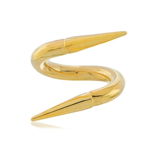 May include: A gold-colored, abstract sculpture with a curved, serpentine design. The piece features two pointed ends and a smooth, polished surface. The sculpture is set against a white background, highlighting its form and metallic sheen.