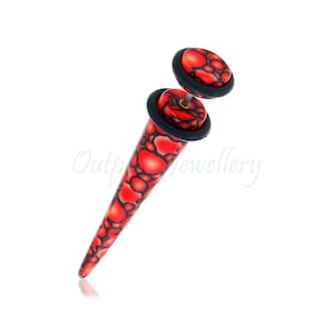 May include: A red and black ear gauge with a tapered design. The gauge features a pattern of red shapes outlined in black. The gauge has a black rubber ring and a matching red and black disc.