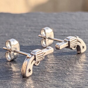 May include: A pair of silver-toned metal stud earrings shaped like miniature revolvers. Each earring features a detailed cylinder, a barrel, and a small star cutout on the handle, complete with a standard post and a circular metal earring back.