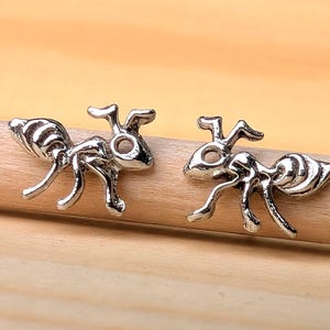 Könnte beinhalten: Ein Paar silberfarbene Ohrstecker in Ameisenform. Die Ohrringe sind mit segmentierten Körpern, Antennen und Beinen versehen. Sie liegen auf einem hellbraunen Holzrundstab.