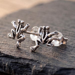 Può includere: Un paio di orecchini a lobo in argento sterling a forma di piccole rane con arti dettagliati e corpi testurizzati. Ogni orecchino presenta un perno con una chiusura in plastica trasparente e metallo con il marchio 925 impresso.