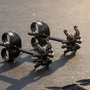 Op de afbeelding: Een paar zwarte spinnen-oorstekers. Elke oorbel heeft een gedetailleerd spinnendesign met acht poten en een kleine pin om door te prikken. De oorbellen zijn geplaatst op een donker, getextureerd oppervlak.