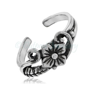 May include: A silver-colored toe ring with a floral design. The ring features a square flower with detailed petals and a central dot. The band has a decorative, swirling pattern and a curved, open design.