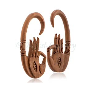 May include: Pair of brown wooden earrings in the shape of hands, each with a curved hook. The hands are carved with intricate details, including fingers and an eye symbol on the palm. The earrings are designed to be worn through stretched ear piercings.