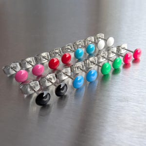 Pode incluir: Uma coleção de brincos de pressão coloridos em várias tonalidades, incluindo rosa, vermelho, azul, verde e preto. Os brincos têm um design simples e redondo com um pino e costas metálicas brilhantes. Os brincos estão dispostos em fileiras sobre uma superfície reflexiva.