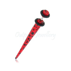May include: A red and black star-patterned ear gauge. The gauge has a conical shape with a flat, circular end and a matching circular disc on the other end. The gauge is designed for stretched ear piercings.