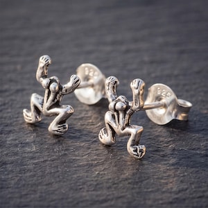May include: A pair of sterling silver stud earrings shaped like stylized frogs in a leaping pose. Each earring features a polished metallic finish with detailed limbs and a stamped 925 hallmark on the circular earring backings.