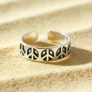 May include: A silver-colored adjustable toe ring featuring a repeating pattern of peace symbols. The ring is open at the top and bottom, and is resting on a bed of sand.