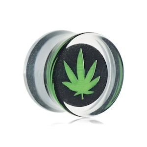 May include: Clear acrylic ear gauge plug with a green cannabis leaf design on a black background. The plug has a rounded shape and is designed for stretched ear piercings. The gauge is visible through the clear acrylic.