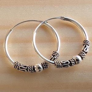 May include: A pair of silver hoop earrings with decorative detailing. The earrings have a circular design with intricate patterns and small silver beads. The earrings are displayed on a light brown wooden surface.