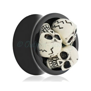 Könnte beinhalten: Schwarzer Ohrstecker mit weißem Totenkopf-Design. Der Stecker hat einen schwarzen, kreisförmigen Rahmen mit einem konkaven Innenraum, der mit drei weißen Totenköpfen gefüllt ist. Die Totenköpfe haben schwarze Augenhöhlen und Rissdetails.