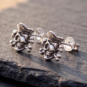 May include: A pair of silver stud earrings shaped like pirate skulls wearing eye patches and hats, with crossed bones behind each skull. The earrings feature a metallic finish and include butterfly backings stamped with the 925 hallmark.