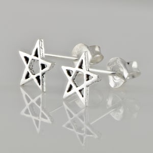 May include: A pair of silver star stud earrings. Each earring has a five-pointed star design with an open centre. The earrings are shown on a reflective surface, highlighting their polished finish and the '925' stamp.