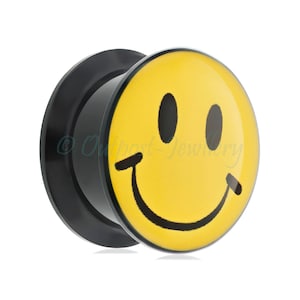 May include: A black ear plug featuring a yellow smiley face. The smiley face has black eyes and a curved black mouth. The product is made of a shiny material and is set against a white background.