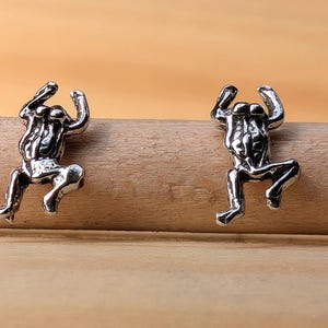 May include: Two silver-colored, stylized human figure stud earrings. The figures are in a climbing pose, with arms raised. The earrings are displayed on a wooden dowel against a light brown background. The earrings are likely made of metal.