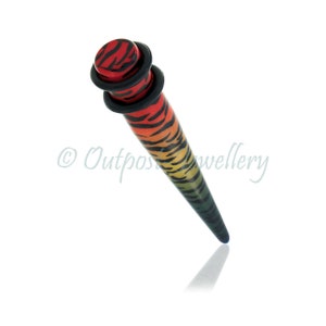 Puede incluir: Un ensanchador de oreja cónico y vibrante con un patrón de rayas de tigre. El ensanchador presenta un degradado de rojo, amarillo y verde, con rayas negras. La parte superior tiene un disco rojo con rayas negras y anillos negros.
