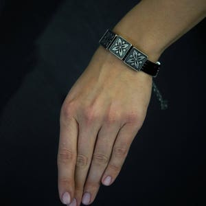 Pode incluir: Um bracelete de couro preto com pingentes quadrados prateados com um design floral.